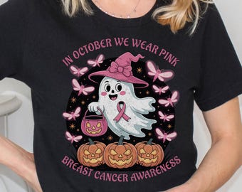 May include: Black t-shirt with a graphic of a ghost in a pink witch hat, holding a pink ribbon, surrounded by pink butterflies and pumpkins. Text reads "IN OCTOBER WE WEAR PINK" and "BREAST CANCER AWARENESS".