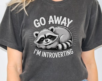 Puede incluir: Camiseta gris oscuro con un gráfico de un mapache durmiendo y el texto "GO AWAY I'M INTROVERTING" en blanco. El mapache es gris y blanco con una máscara negra y cola a rayas. Estilo casual.