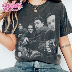 May include: A charcoal gray short-sleeve t-shirt featuring a black and white vintage-style photographic print of four individuals. The graphic shows the group in a candid arrangement, with one person wearing a jersey with visible text on the sleeve.