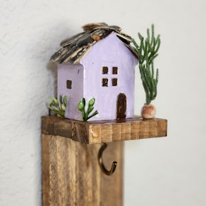 May include: A miniature lavender house with a seed-covered roof sits on a small wooden shelf with a hook. The house has brown windows and a door, with small green plants and a tree-like decoration adding detail.