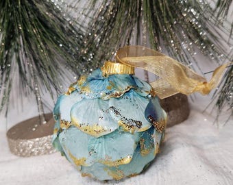 May include: A festive Christmas bauble with a blue and gold petal design. The bauble features a gold cap and a gold ribbon for hanging. The petals are light blue with gold edges and black glitter accents. It's set against a backdrop of pine needles.