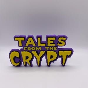 Puede incluir: Logotipo morado y amarillo de "Tales From The Crypt". El texto está en negrita, con un estilo de dibujos animados, con relleno amarillo y contorno morado. El fondo es de color morado claro.