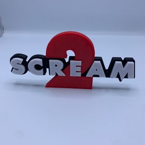 Könnte beinhalten: Ein 3D-gedrucktes Schild mit der Aufschrift "SCREAM 2" in schwarz-weißen Buchstaben mit einer roten Zahl 2.