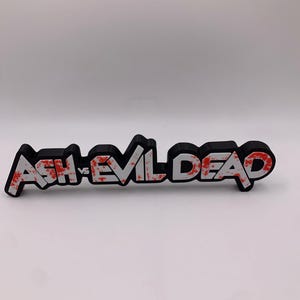 Puede incluir: Señal impresa en 3D en blanco y negro con las palabras "Ash vs Evil Dead" en salpicaduras de pintura roja y blanca.