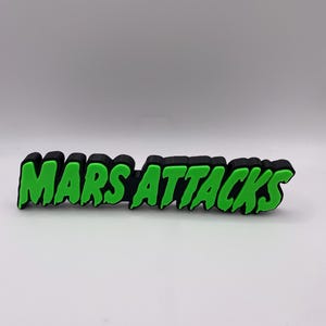 以下が含まれることがあります： 黒と緑の「MARS ATTACKS」ロゴ。テキストは太字で、緑色の文字と黒いアウトラインで表示されています。ロゴは長方形です。