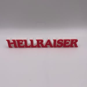 Peut inclure: Un panneau blanc et rouge avec l'inscription "HELLRAISER". Les lettres sont blanches avec des contours et des dessus rouges. Le panneau est posé sur une surface blanche.