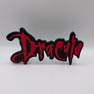 Puede incluir: Un emblema negro y rojo con la palabra "Drácula". La palabra está escrita en una fuente roja goteante, con un contorno negro. Probablemente sea un artículo decorativo o un accesorio.