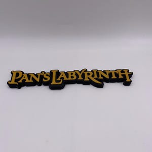 Puede incluir: Un letrero negro y dorado con el texto "Pan's Labyrinth". El texto está en una fuente dorada estilizada con un contorno negro. Es probable que sea un artículo decorativo o de merchandising.
