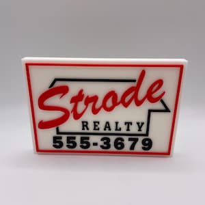 May include: A rectangular sign with a white background and a red border. The sign features the name "Strode" in red cursive, with "REALTY" and a phone number in black. The sign is likely for a real estate business.