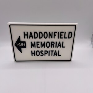 Könnte beinhalten: Ein weißes Schild mit schwarzem Text und schwarzem Rand. Auf dem Schild steht "HADDONFIELD MEMORIAL HOSPITAL" mit einem nach links weisenden Pfeil. Die Buchstaben "HMH" befinden sich im Pfeil. Das Schild ist rechteckig mit einer leichten Trapezform.