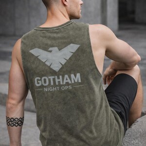 May include: Olive green tank top with a gray bat symbol and the text "GOTHAM NIGHT OPS." The sleeveless shirt is made of a distressed fabric. The person in the image is wearing black shorts and has a tattoo on their arm.
