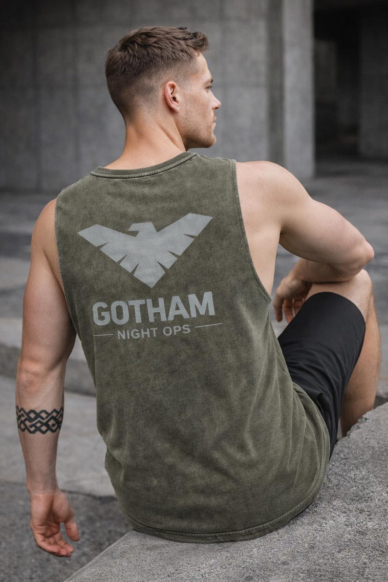 May include: Olive green tank top with a gray bat symbol and the text "GOTHAM NIGHT OPS." The sleeveless shirt is made of a distressed fabric. The person in the image is wearing black shorts and has a tattoo on their arm.