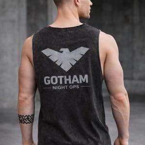 May include: Dark gray tank top with a distressed finish. The back features a silver graphic of a stylized bird above the text "GOTHAM NIGHT OPS". The sleeveless shirt is made of a soft material.