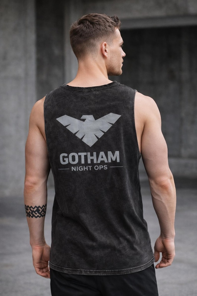 May include: Dark gray tank top with a distressed finish. The back features a silver graphic of a stylized bird above the text "GOTHAM NIGHT OPS". The sleeveless shirt is made of a soft material.