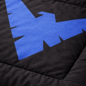 Może przedstawiać: Czarna pikowana tkanina z dużym niebieskim geometrycznym wzorem. Wz&oacute;r to stylizowany kształt ptaka lub nietoperza. Tkanina ma miękki, teksturowany wygląd z widocznymi szwami.