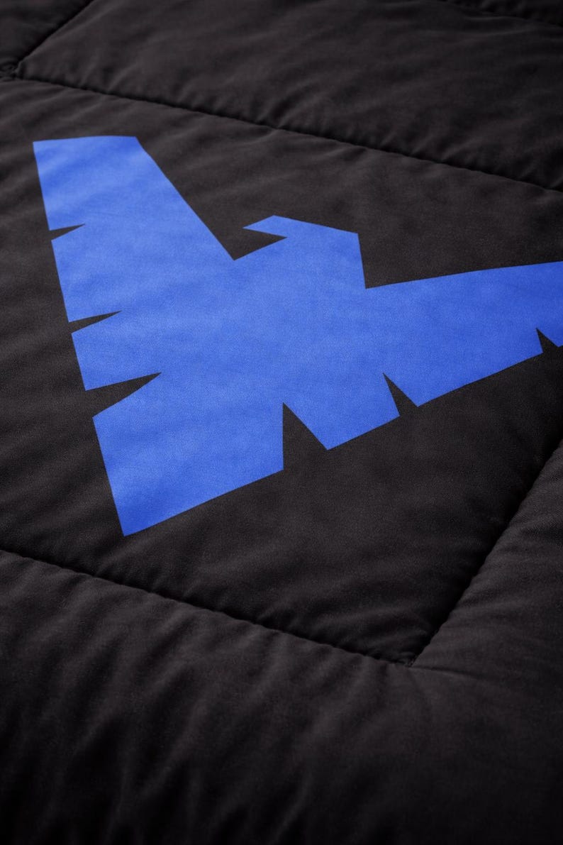 Może przedstawiać: Czarna pikowana tkanina z dużym niebieskim geometrycznym wzorem. Wz&oacute;r to stylizowany kształt ptaka lub nietoperza. Tkanina ma miękki, teksturowany wygląd z widocznymi szwami.