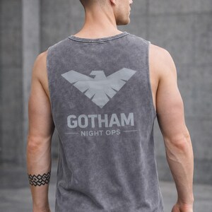 May include: Gray tank top with the text "GOTHAM NIGHT OPS" and a stylized bird emblem on the back. The sleeveless shirt is made of a soft, textured fabric.