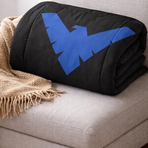 Może przedstawiać: Złożony czarny koc z dużym, stylizowanym niebieskim emblematem ptaka. Koc spoczywa na jasno szarym tapicerowanym krześle, z beżowym kocem z frędzlami widocznym z boku.