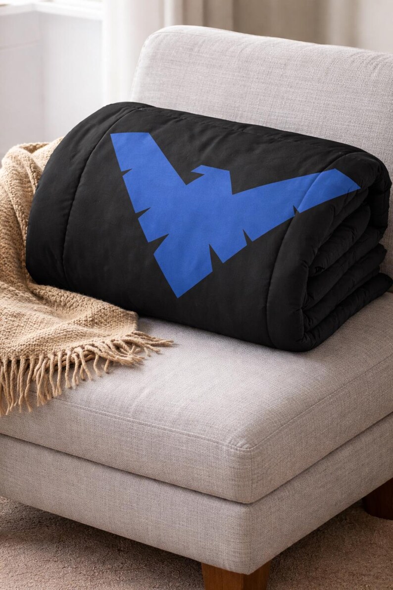 Może przedstawiać: Złożony czarny koc z dużym, stylizowanym niebieskim emblematem ptaka. Koc spoczywa na jasno szarym tapicerowanym krześle, z beżowym kocem z frędzlami widocznym z boku.