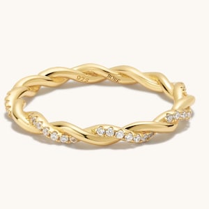 以下が含まれることがあります： 縁に小さな透明な石がセットされた、ゴールドのツイストバンドリング。リングには「JIORE」と「D25」の刻印があります。