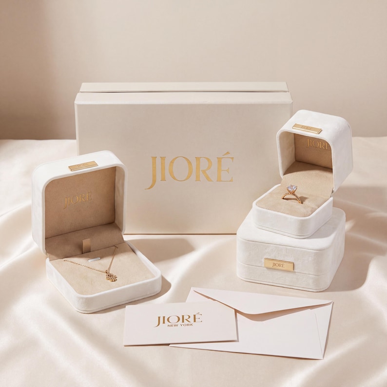 K&ouml;nnte beinhalten: Eine Kollektion cremefarbener Schmuckschatullen mit goldenem JIOR&Eacute;-Logo, die eine goldene Halskette mit Hamsa-Anh&auml;nger und einen goldenen Ring mit klarem Stein enthalten. Ein passender cremefarbener Umschlag mit JIOR&Eacute; NEW YORK-Logo liegt davor.