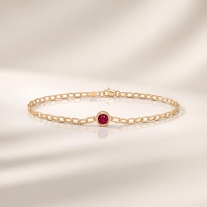 Könnte beinhalten: Ein goldenes Kettenarmband mit einem einzelnen runden, in einer Zargenfassung gefassten Rubin in der Mitte. Das Armband besteht aus gleichmäßigen ovalen Gliedern und verfügt über einen kleinen Karabinerverschluss auf der Rückseite.