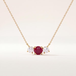 May include: A delicate gold chain necklace featuring a central round-cut deep red ruby flanked by two smaller round-cut clear white diamonds. The stones are set in a simple gold prong setting, creating a classic three-stone pendant design.