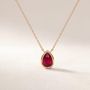 May include: A gold chain necklace featuring a single pear-shaped, faceted red gemstone pendant set in a smooth gold bezel. The vibrant red stone is suspended from a delicate gold link chain, creating a simple and elegant piece of jewelry.