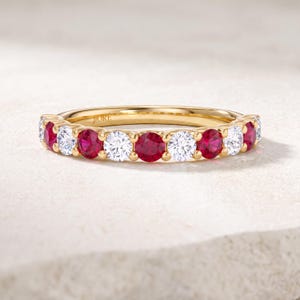 May include: A gold band ring featuring an alternating pattern of round-cut red rubies and clear white diamonds set in a shared prong setting. The interior of the gold band is engraved with the brand name JIORE in a clear, legible font.