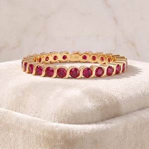 May include: A gold eternity band featuring a continuous row of round, vibrant red rubies set in a polished metal frame. The ring displays a repeating S-shaped design between each gemstone, with the brand name JIORÉ engraved on the inner band.
