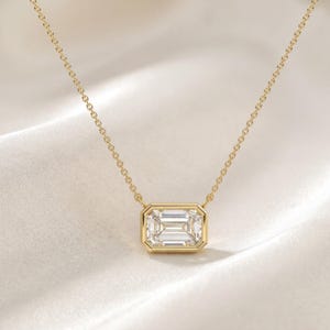 May include: A gold necklace featuring a rectangular emerald-cut clear gemstone set in a polished gold bezel setting. The pendant is suspended from a delicate gold chain with fine links, creating a clean and symmetrical design for the jewelry piece.