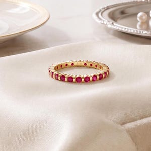 K&ouml;nnte beinhalten: Ein goldener Eternity-Ring mit einer durchgehenden Reihe runder, leuchtend pink-roter Rubine in Kanalfassung. Die Innenseite des Rings ist mit dem Text JIORE graviert. Der Ring ist kreisf&ouml;rmig und hat eine polierte, metallische Oberfl&auml;che.