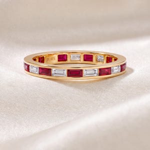 May include: A gold band ring featuring an alternating pattern of baguette-cut red rubies and clear white diamonds. The interior of the band is engraved with the text JIORÉ. The stones are set in a channel style around the entire circumference.