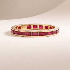 Op de afbeelding: Een gouden ring met een doorlopende rij rechthoekige, baguette-geslepen rode edelstenen in een kanaalzetting. De binnenzijde van de gouden ring is gegraveerd met de merknaam JIORÉ in een duidelijk, schreefloos lettertype.