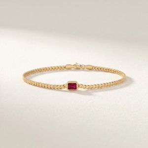 Può includere: Un bracciale a catena in oro con una singola pietra preziosa rossa rettangolare a taglio smeraldo, incastonata in una cornice a lunetta al centro. Il bracciale è composto da maglie a catena intrecciate e una chiusura a moschettone.