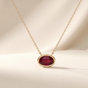 May include: A gold necklace featuring an oval-shaped, deep red ruby gemstone set in a smooth, polished gold bezel setting. The pendant is suspended from a delicate gold chain with small, uniform links that connect to the sides of the oval frame.