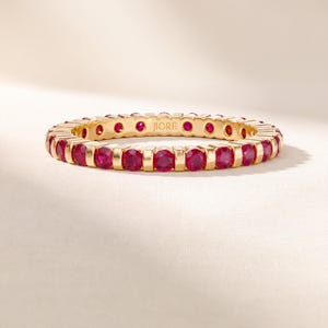 May include: A gold eternity band featuring a continuous row of round, vibrant red rubies set in a polished metal frame. The interior of the band is engraved with the brand name JIORÉ in a clear, sans-serif font, showcasing a refined design.