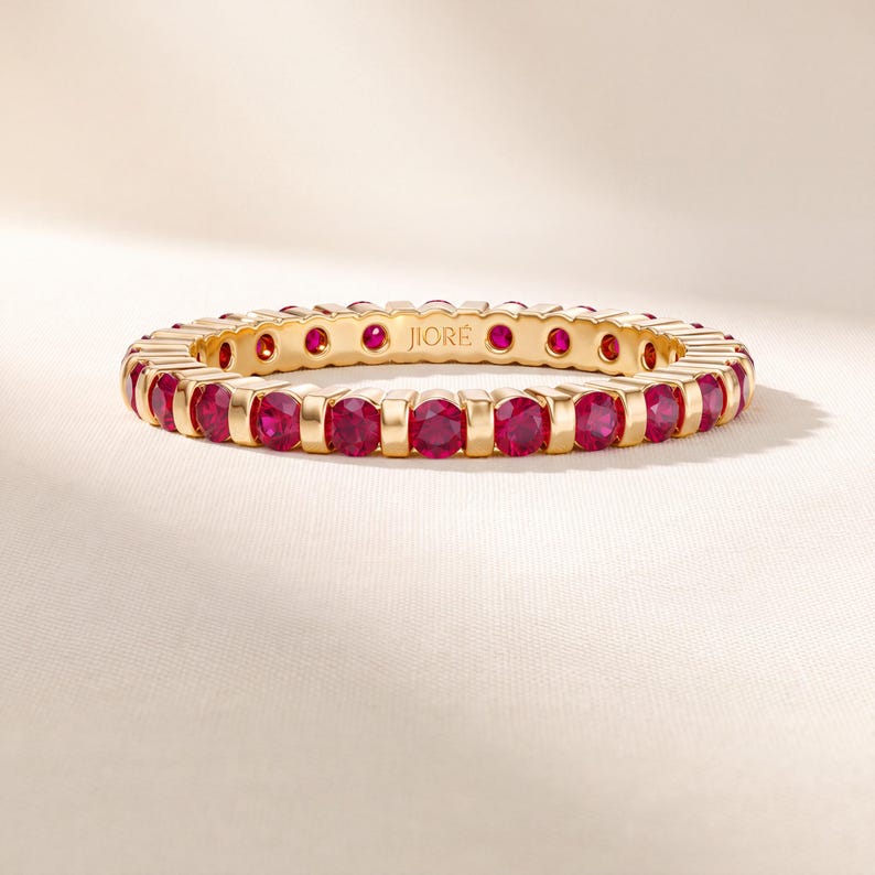K&ouml;nnte beinhalten: Ein goldener Eternity-Ring mit einer durchgehenden Reihe runder, leuchtend roter Rubine, die in einen polierten Metallrahmen gefasst sind. Die Innenseite des Rings ist mit dem Markennamen JIOR&Eacute; in einer klaren serifenlosen Schrift graviert.