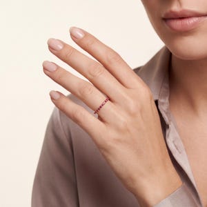 K&ouml;nnte beinhalten: Ein d&uuml;nner, zierlicher Ring mit einem durchgehenden Band aus kleinen, leuchtend roten Edelsteinen, die in einen polierten Metallrahmen gefasst sind. Der Ring wird am Ringfinger getragen und zeigt ein schlichtes Design mit einer gleichm&auml;&szlig;igen Reihe.