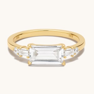 May include: A gold ring with a rectangular emerald-cut diamond and two smaller baguette-cut diamonds on either side.