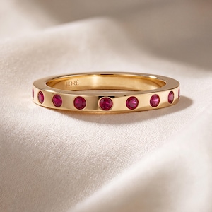 May include: A gold band ring featuring a series of flush-set, round, vibrant red rubies encircling the exterior. The interior of the band is engraved with the text JIORÉ. The ring has a polished finish and a consistent, slim, circular profile.