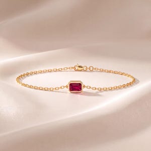 May include: A delicate gold chain bracelet featuring a single rectangular emerald-cut ruby gemstone set in a polished gold bezel. The bracelet includes a small lobster claw clasp mechanism at the back, showcasing a refined and minimalist jewellery design.