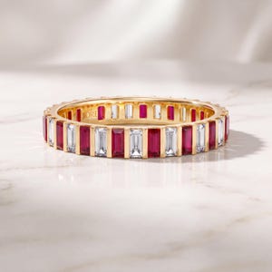 May include: A gold eternity band featuring alternating baguette-cut red rubies and clear white diamonds set in a channel setting. The interior of the gold band is engraved with the brand name JIORE in a clear, legible font.