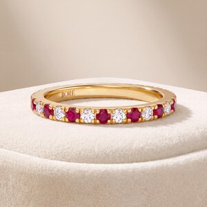 May include: A yellow gold eternity band featuring an alternating pattern of round-cut red rubies and clear white diamonds set in a shared prong setting. The interior of the band is engraved with the brand name JIORÉ in a clean, sans-serif font.