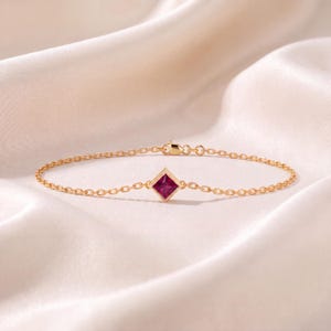 May include: A delicate gold chain bracelet featuring a single square-cut, deep pink gemstone set in a gold frame at the center. The bracelet includes a small lobster claw clasp and thin, polished gold links that connect to the central stone.