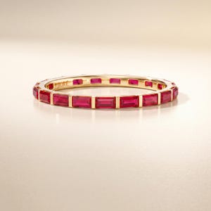 May include: A gold eternity band featuring a continuous row of baguette-cut red rubies set in a channel. The interior of the ring is engraved with the brand name JIORÉ in a serif font. The metal has a polished finish and a warm yellow tone.