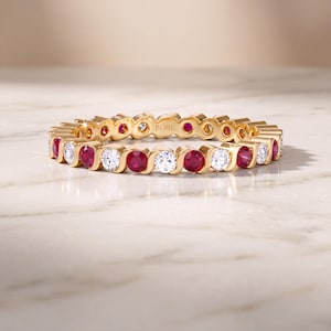 Puede incluir: Una alianza de oro amarillo con un patrón alterno de rubíes rojos y diamantes blancos de talla redonda, engastados en un diseño metálico ondulado continuo. El interior del anillo tiene grabado el nombre de la marca JIORE en mayúsculas.