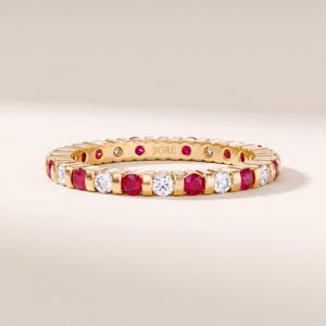 May include: A gold eternity band featuring an alternating pattern of round-cut red rubies and clear white diamonds set in a continuous row. The interior of the band is engraved with the brand name JIORÉ in a clear, capitalized font.