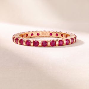 K&ouml;nnte beinhalten: Ein Ewigkeitsring aus Ros&eacute;gold mit einer durchgehenden Reihe runder, leuchtend pink-roter Rubine in individuellen Metallfassungen. Die Innenseite des Rings ist deutlich mit dem Markennamen JIORE in einer serifenlosen Schrift graviert.