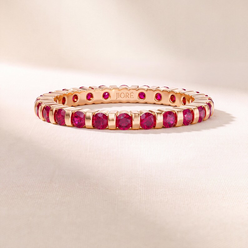 K&ouml;nnte beinhalten: Ein Ewigkeitsring aus Ros&eacute;gold mit einer durchgehenden Reihe runder, leuchtend pink-roter Rubine in individuellen Metallfassungen. Die Innenseite des Rings ist deutlich mit dem Markennamen JIORE in einer serifenlosen Schrift graviert.